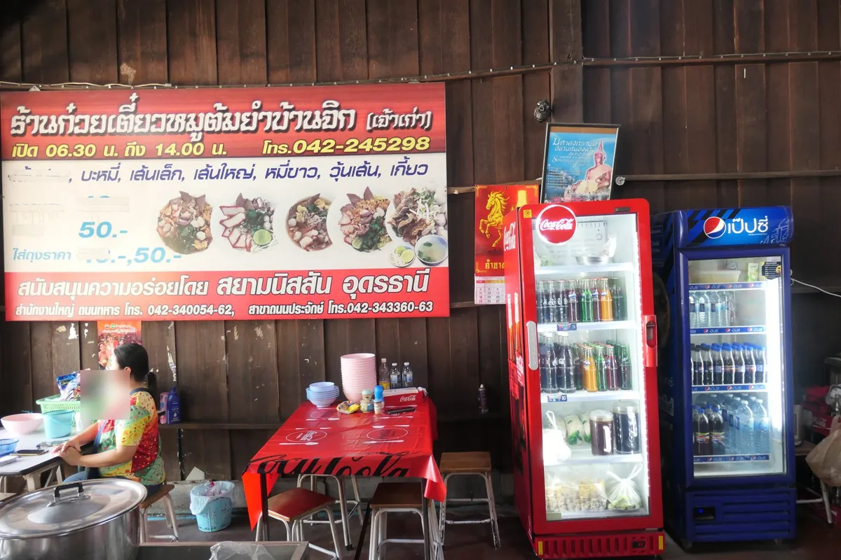 ป้ายเมนูสีแดงบนผนังไม้ โต๊ะและตู้เย็นน้ำอัดลมในร้าน มีแม่ค้านั่งอยู่หน้าหม้อต้ม