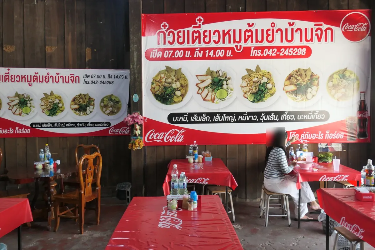โต๊ะพลาสติกสีแดงและเก้าอี้ไม้ในร้านติดผนังไม้ มีป้ายเมนูใหญ่สีขาวแดง