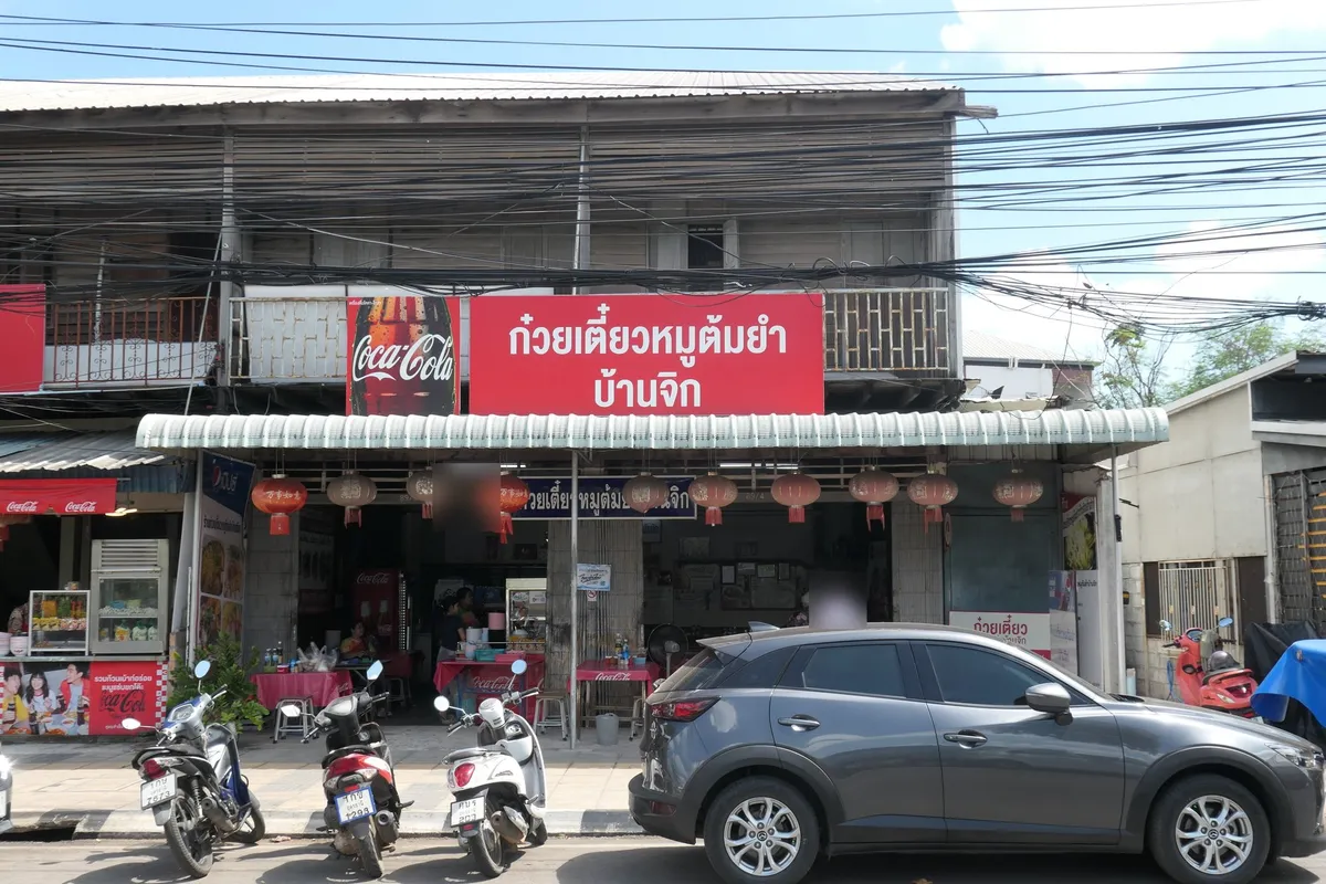 ร้านก๋วยเตี๋ยวหมูต้มยำบ้านจิกหน้าตัวอาคารไม้สองชั้น ป้ายสีแดงตัวอักษรขาว มีโต๊ะและรถยนต์จอดอยู่หน้าร้าน