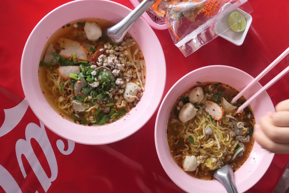 ป้ายเมนูก๋วยเตี๋ยวหมูต้มยำบ้านจิกบนผนังไม้ มีรูปชามก๋วยเตี๋ยวหลายแบบ ราคาเริ่มต้น 50 บาท และตู้น้ำอัดลมอยู่ข้างๆ