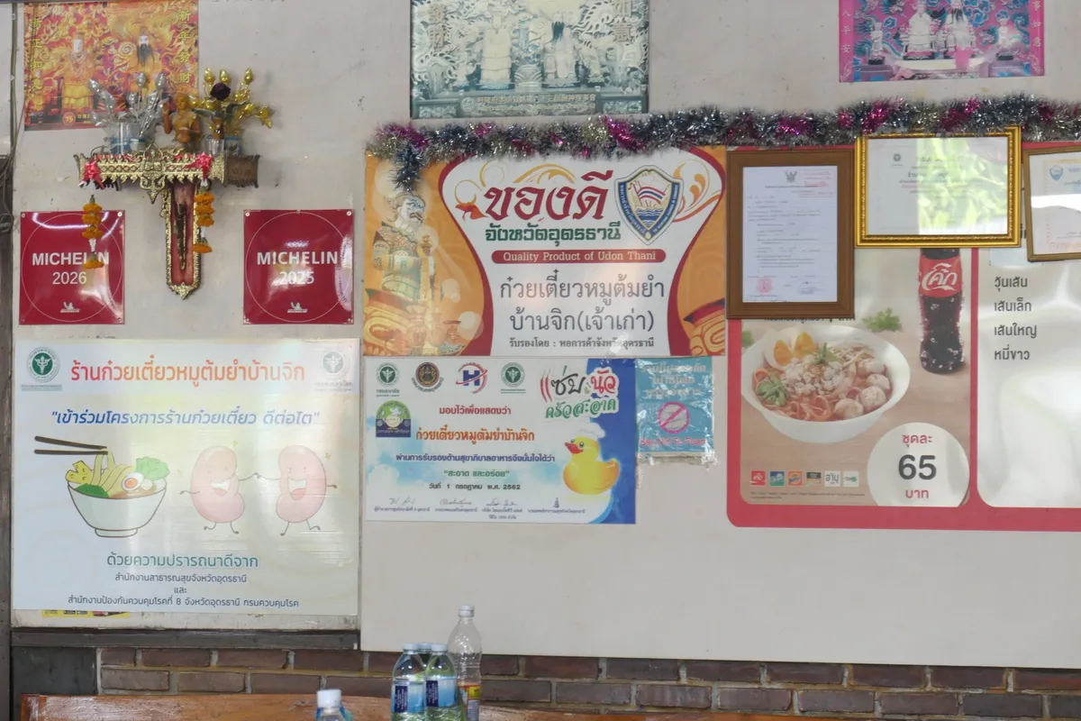 ป้ายสีแดงสองใบ เขียนว่า MICHELIN 2026 และ MICHELIN 2025 ติดอยู่บนผนังร้าน ใกล้ป้ายของดีจังหวัดอุดรธานี