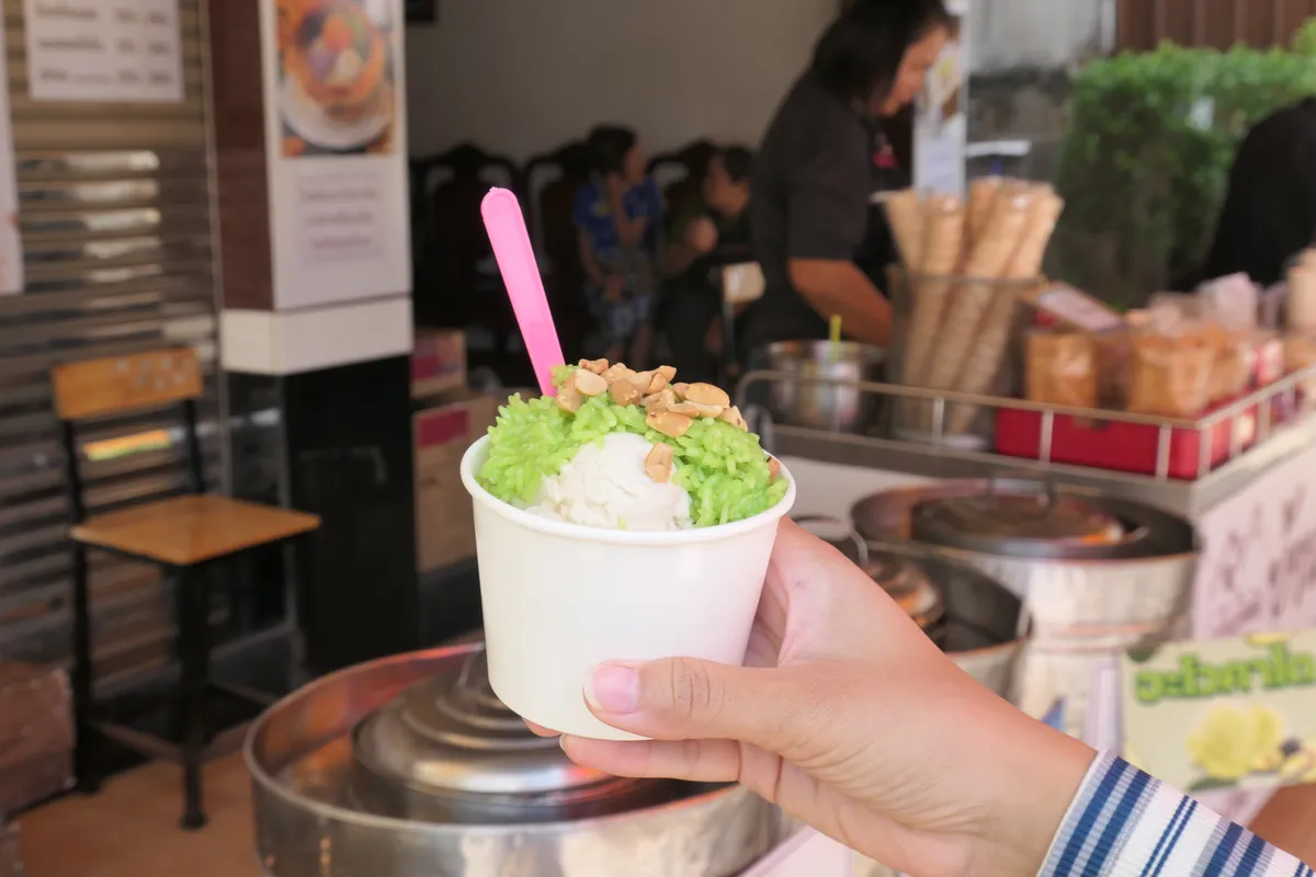 A cup of coconut ice cream on green pandan sticky rice topped with peanuts, held in a white paper cup, with the shop blurred in the background