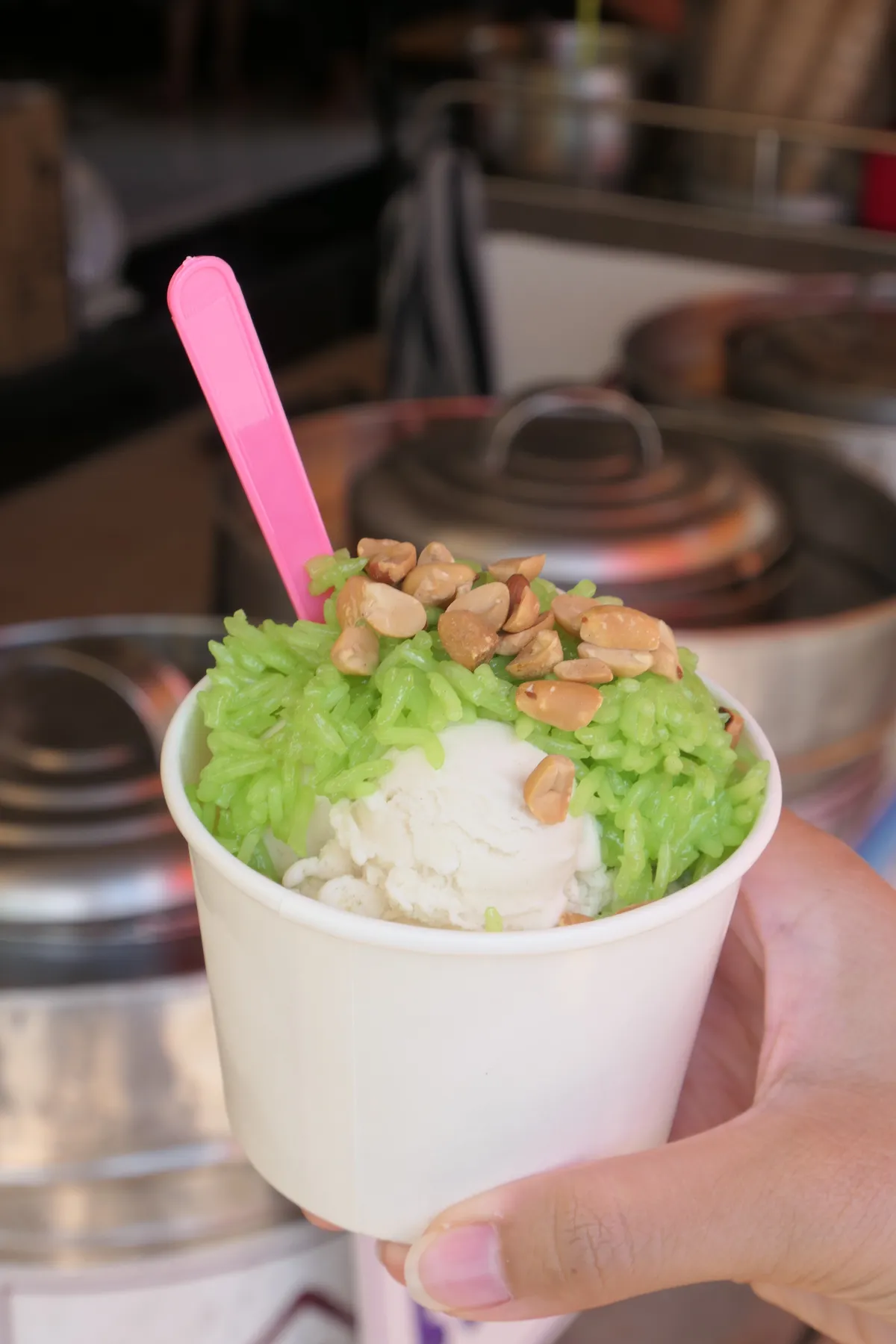 Coconut ice cream with green pandan sticky rice and peanuts in a paper cup, with a pink plastic spoon