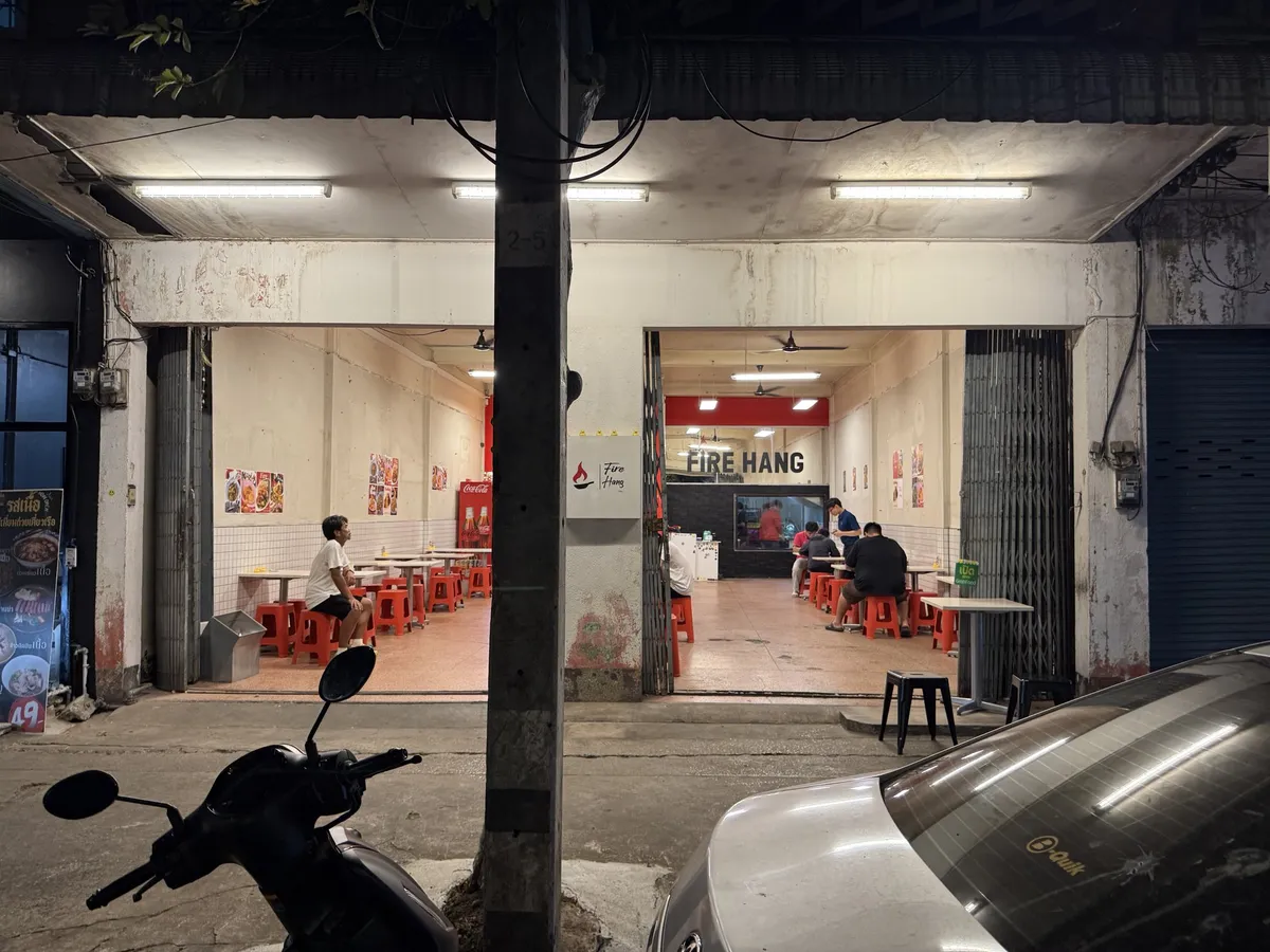 หน้าร้านไฟแฮงสุกี้ตอนกลางคืน มีแสงไฟจากร้านและโต๊ะเก้าอี้สีแดงด้านใน มีมอเตอร์ไซค์จอดหน้าร้าน