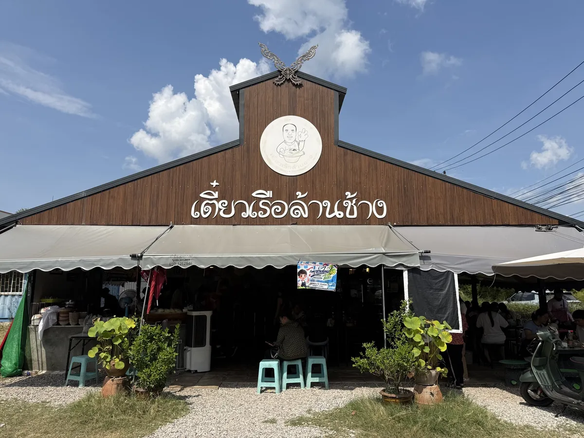 หน้าร้านเตี๋ยวเรือล้านช้าง อาคารไม้สีน้ำตาลมีป้ายชื่อร้านสีขาวอยู่ด้านบน มีเก้าอี้พลาสติกวางหน้าร้าน