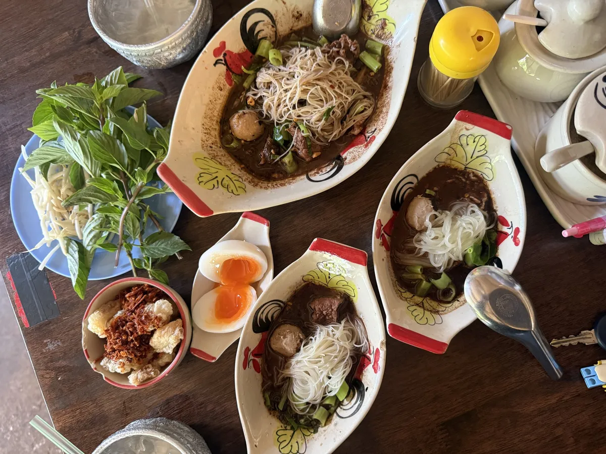 เตี๋ยวเรือสองชามในถ้วยเรือสีขาวลายดอก มีเส้น ลูกชิ้น ไข่ต้ม และผักสด วางบนโต๊ะไม้