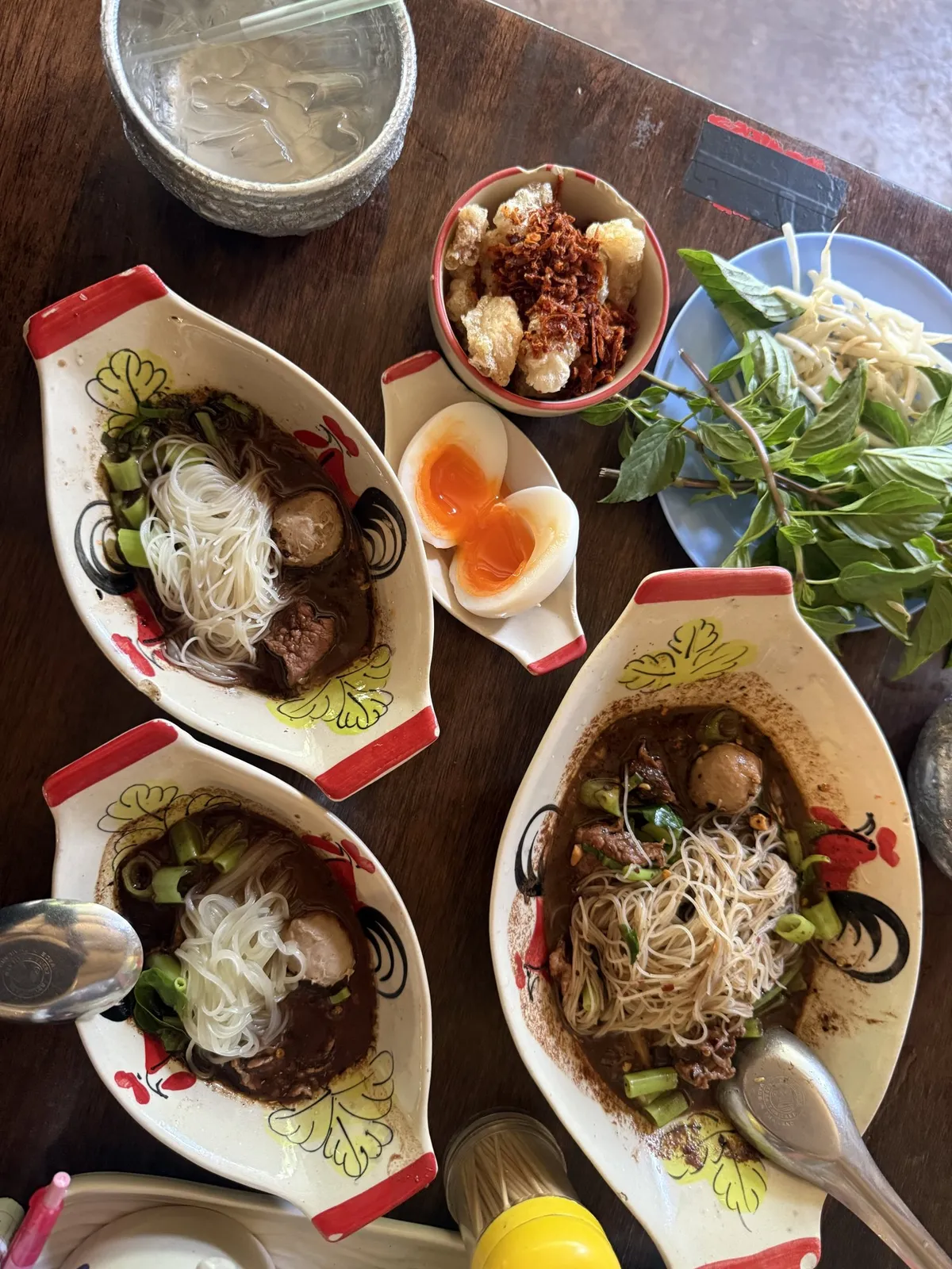 ชามเตี๋ยวเรือหลายชามในถ้วยเรือลายดอก ไข่ต้มครึ่งลูก เครื่องเคียงผักสด และน้ำจิ้ม