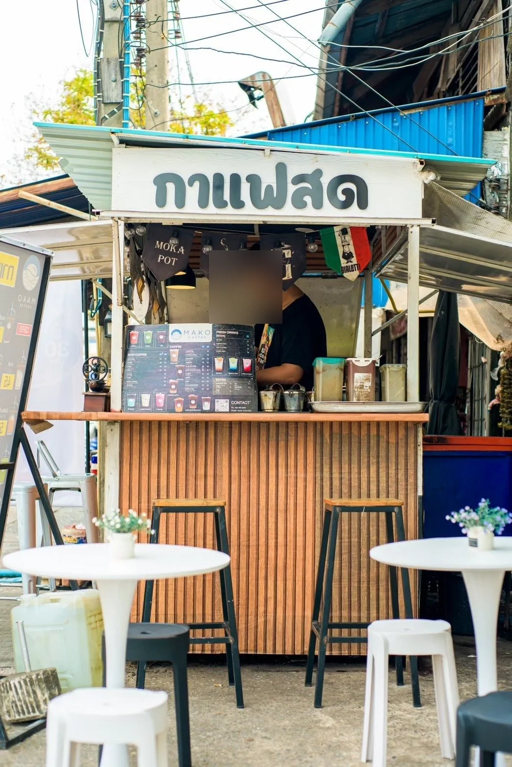 เคาน์เตอร์กาแฟสดสีส้ม ป้ายกาแฟสดด้านบน มีโต๊ะกลมขาวและเก้าอี้บาร์สำหรับนั่งรอ