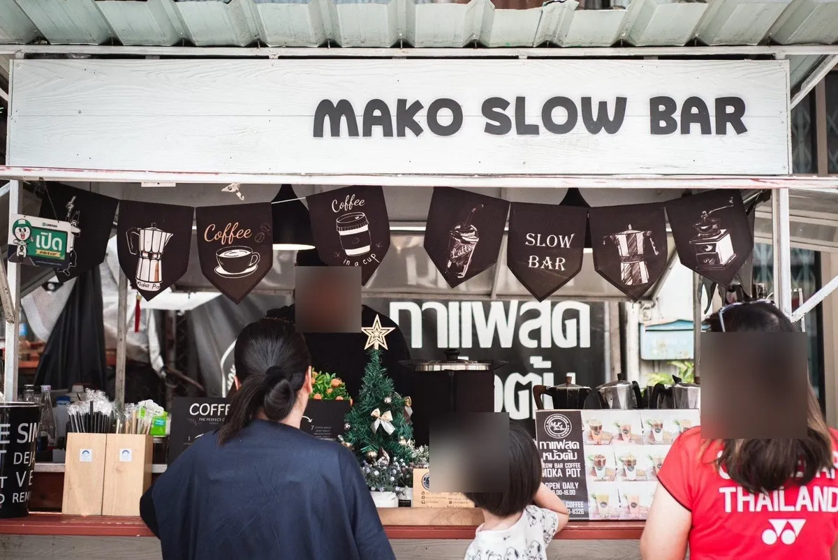 ร้านกาแฟเคาน์เตอร์ไม้สีส้ม มีป้ายกาแฟสดและโต๊ะกลมขาวกับเก้าอี้บาร์ด้านหน้า