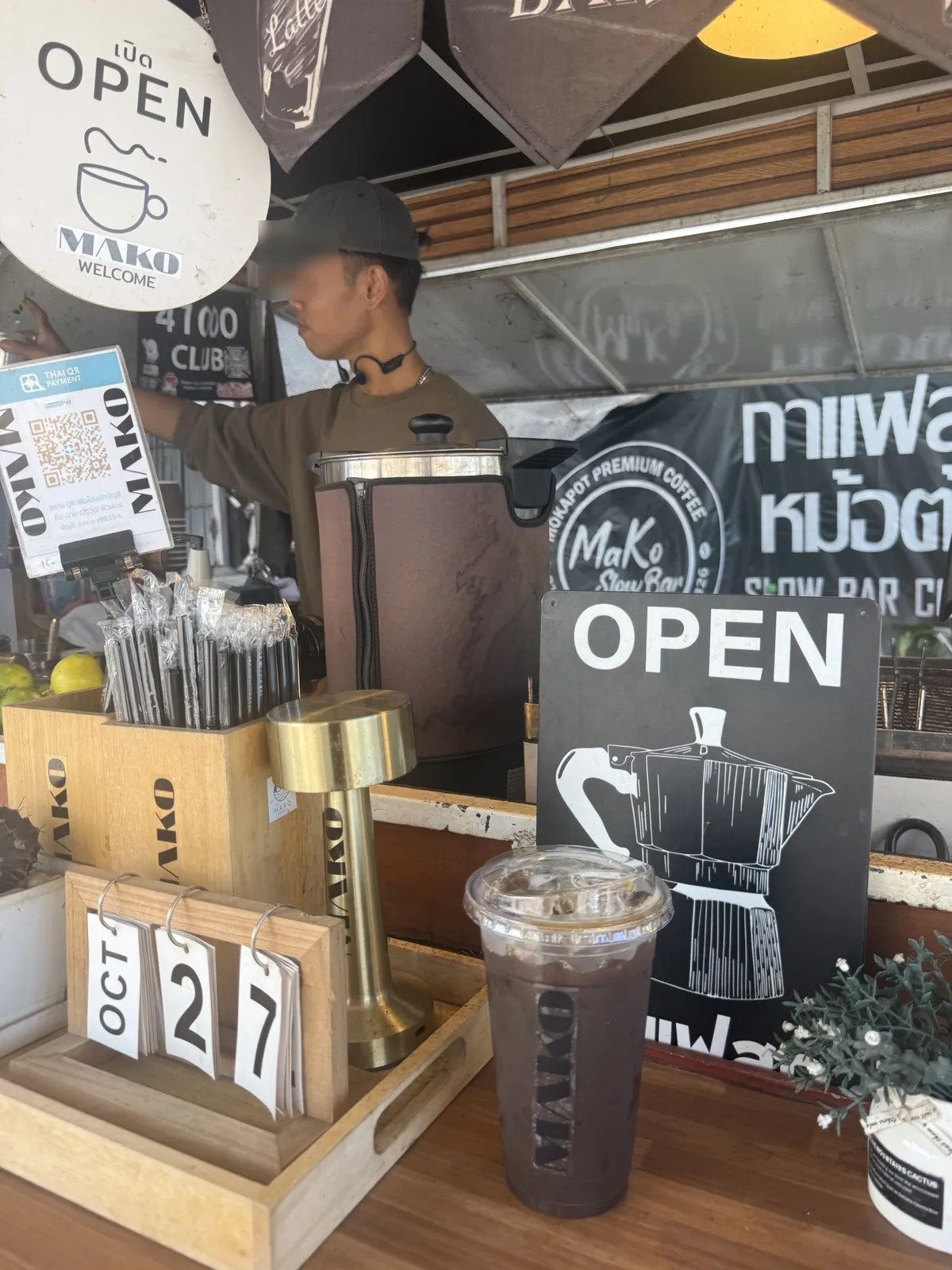 เคาน์เตอร์ร้านมาโกะ มีป้าย OPEN และหม้อ Moka Pot ภาพวาด แก้วกาแฟเย็นสีเข้มมีโลโก้ MAKO วางอยู่บนโต๊ะไม้